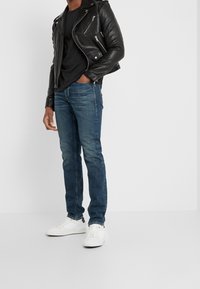 Chaqueta de motociclista de cuero negro, camiseta negra ajustada, jeans de mezclilla azul y zapatillas blancas, de pie sobre un fondo neutro.