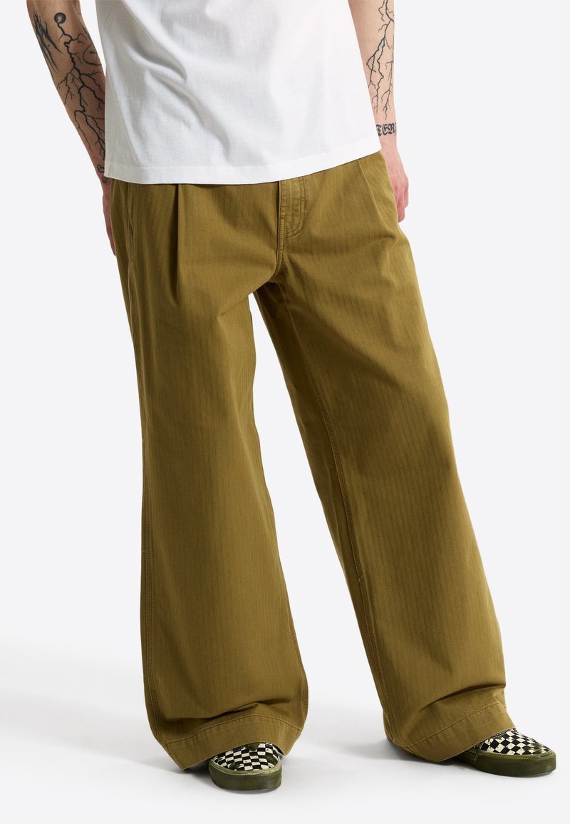 Persona que lleva pantalones sueltos de color verde oliva, camiseta blanca y zapatos tipo slip-on a cuadros en blanco y negro, de pie con los brazos tatuados visibles.
