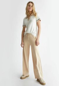 Polo shirt bianco a maniche corte con colletto, abbinato a pantaloni beige a gamba larga e sneakers beige con suola spessa. Tessuto della stoffa liscio.