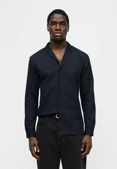 Άνδρας με σκούρο μπλε πουκάμισο με κουμπιά και γιακά και μαύρο τζιν, που στέκεται μπροστά, με φόντο απλό λευκό.