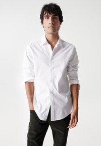 Camisa blanca de botones con un cuello nítido, llevada con las mangas remangadas. El modelo la combina con pantalones oscuros, presentando un look limpio y clásico.