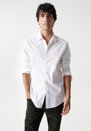 Camisa blanca de botones con un cuello nítido, llevada con las mangas remangadas. El modelo la combina con pantalones oscuros, presentando un look limpio y clásico.
