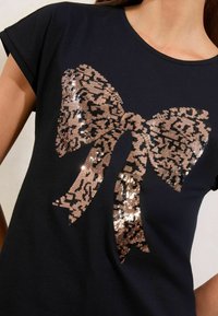 T-shirt nera con un grande motivo a fiocco in paillettes rosa oro con un pattern leopardato. Maniche corte e scollatura rotonda.