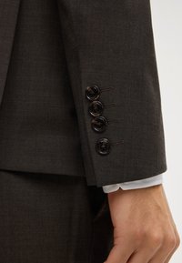 Manica di un abito marrone con quattro bottoni scuri e un polsino di camicia bianca visibile, accanto a una parte di una mano e pantaloni marroni coordinati.