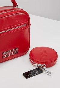 Czerwona torba Versace Jeans Couture i okrągły woreczek na monety ze srebrnymi zamkami błyskawicznymi i plakietkami z logo na białej powierzchni.