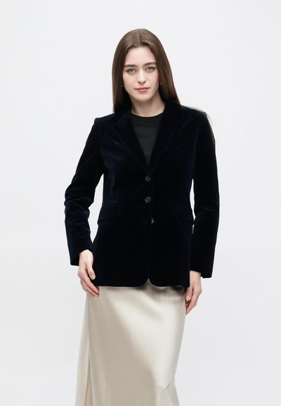Blazer di velluto blu navy con revers a lancia, due tasche frontali e tre bottoni. Abbinato a una gonna di raso beige.