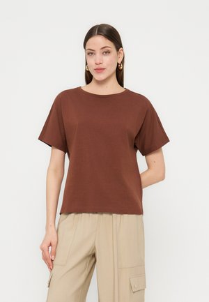 Žena s rovnými tmavými vlasmi má na sebe hnedý krátky top, béžové cargo nohavice a zlaté kruhové náušnice, pózuje na jednoduchom pozadí.