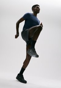 Czarne sportowe buty z systemem sznurowania, siateczkową cholewką i gumową podeszwą. Model nosi niebieską koszulkę i szare szorty, przyjął pozę z uniesionym kolanem.