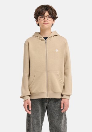 Adolescent garçon aux cheveux bruns et lunettes portant un sweat à capuche beige zippé et un jean gris, debout devant un fond uni.