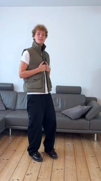 Zöld párnázott mellény fehér póló felett, fekete bő nadrág és fekete cipő, fából készült padlón áll egy világos, jól megvilágított szobában.