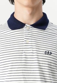 Bela in mornarsko modra polo majica s črtami, mornarsko modrim ovratnikom, dvema gumboma in vezenim logotipom "GAP" na levem prsnem delu. Bombažna struktura.