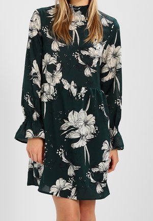 Kvinde iført en mørkegrøn langærmet kjole med store hvide blomsterprint og flæsekant ved ærmerne, stående mod en ensfarvet baggrund.