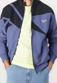 Reebok windbreaker jacket in purple and black, featuring geometric design, zip closure, elastic cuffs, and smooth texture. Worn over a light green shirt.