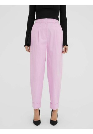 Kobieta ubrana w jasnoróżowe spodnie z wysokim stanem i plisami, z podwiniętymi nogawkami, czarną bluzkę z długim rękawem oraz czarne szpilki z ostrym noskiem, stojąca.