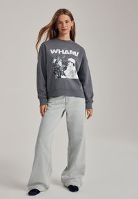 Grauer Pullover mit "WHAM!"-Grafik, locker geschnitten, mit Rippbündchen, kombiniert mit hellgrauen Jeans mit weitem Bein und schwarzen Slip-On-Schuhen.
