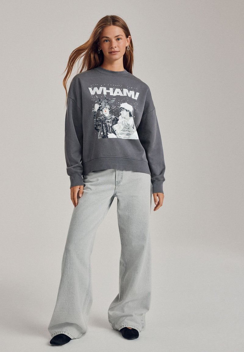 Grauer Pullover mit "WHAM!"-Grafik, locker geschnitten, mit Rippbündchen, kombiniert mit hellgrauen Jeans mit weitem Bein und schwarzen Slip-On-Schuhen.