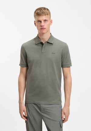 Giovane uomo che indossa una polo verde tenue con il logo "BOSS" sul petto e pantaloni cargo coordinati, in piedi davanti a uno sfondo bianco neutro.