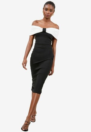 Černé upnuté šaty s bílým off-the-shoulder mašlovým detailem, ke kolenům, hladká textura, jemné řasení, doplněné černými sandály na tenkých popruzích.