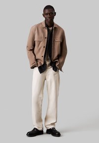 Chaqueta ligera color canela con dos bolsillos frontales, sobre una camisa oscura, combinada con pantalones cremosos de corte holgado y zapatos negros.