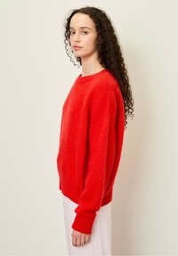 Raudonas, per didelis megztinis su apvalia apykakle ir banguotais rankogaliais, pasižymintis minkšta tekstūra. Modelis jį derina su šviesios spalvos apatine dalimi.