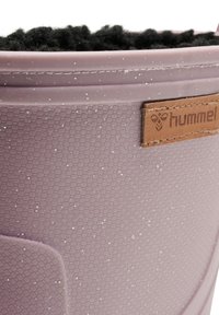 Botte en caoutchouc violet avec un extérieur texturé, doublure en polaire noire et une étiquette en cuir marron avec un logo en relief. Détails subtils en paillettes présents.