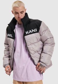 Pufová bunda s čiernou hornou časťou a šedou dolnou časťou. Obsahuje vyšívané texty "KANI" a "KARL." Zapínanie na zips a rebrované manžety.