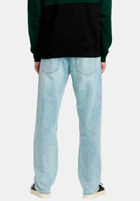 Persona vestida con jeans rectos de color azul claro, sudadera negra y verde, y zapatillas negras con suelas blancas, de pie de espaldas.