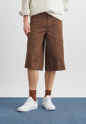 Persona vestida con pantalones cortos marrones hasta la rodilla, calcetines marrones, zapatillas blancas, una camiseta blanca y una camisa de manga larga a cuadros remangada, de pie sobre una alfombra azul.