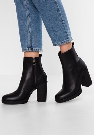 Par sorte læder ankelstøvler med høje hæle og lynlås i siden, båret med cropped blå denim jeans mod en ensfarvet baggrund.
