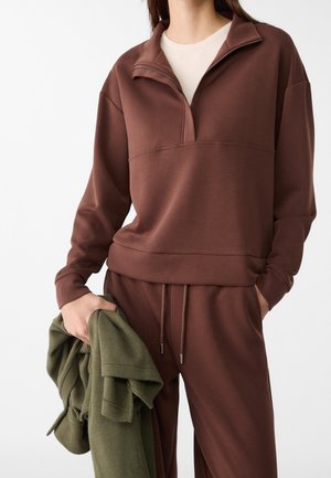 Persoon draagt een bruine zip-up trui en bijpassende broek, houdt een opgevouwen groene fleecejas in één hand vast.