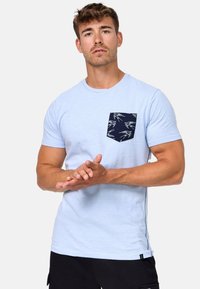 Camiseta de algodón azul claro con un bolsillo azul marino que presenta un patrón de pájaros blancos. Tiene mangas cortas y un escote redondo. Diseño simple y casual.