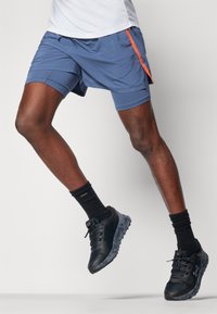 Pantalones cortos azul para hombre con cintura elástica y detalles laterales naranjas, combinados con zapatillas deportivas negras y calcetines negros hasta el tobillo.