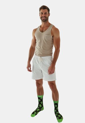 Beige tank top, white shorts, and green alien-patterned socks with black background. Model stands smiling with hands on hips.