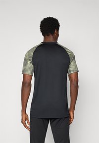 Camiseta deportiva de manga corta con cuerpo negro y mangas raglán de un patrón amarillo-verde, que presenta una textura suave y un cuello redondo.