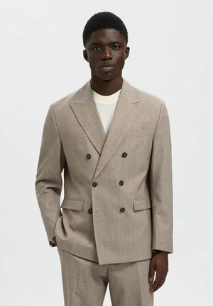 Man wearing beige double-breasted suit with black buttons and white crew neck shirt, standing against plain light gray background.