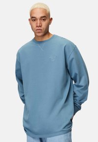Blå sweatshirt i mjukt tyg, med rund halsringning och långa ärmar. Inkluderar en diskret logotyp på vänster bröst och ribbade muddar.