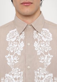 Camicia a righe beige con ricami floreali bianchi al centro, dotata di colletto a punta e chiusura con bottoni. Tessuto testurizzato.