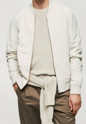 Mężczyzna w jasnobeżowej kurtce bomberce na beżowej koszuli, z beżowym swetrem związanym w talii i brązowych spodniach, z rękami częściowo w kieszeniach.