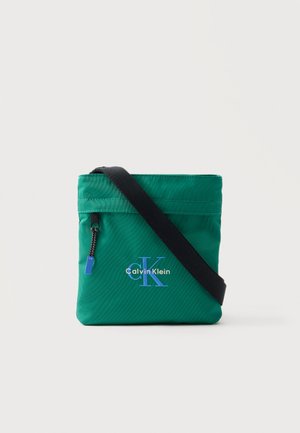 Zelena torbica čez telo s črnim trakom, sprednjim žepom z zadrgo in modrim logotipom "Calvin Klein" na enobarvnem ozadju.