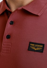 Burgundska polo majica z teksturirano tkanino, tremi črnimi gumbi ter črno oznako z zlatim napisom in obliko kril na prsih.
