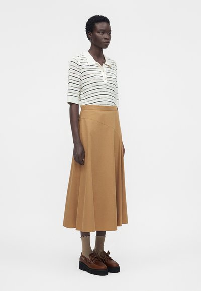 Γυναίκα στέκεται φορόντας λευκό ριγωτό πλεκτό μπλουζάκι με κοντά μανίκια, midi φούστα καμηλό, μπεζ κάλτσες και καφέ παπούτσια με πλατφόρμα σε λευκό φόντο.