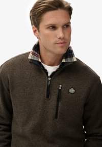 Pullover in pile marrone con collo zip, dotato di fodera a quadri e un patch con logo sul petto. Design testurizzato, caldo e informale.