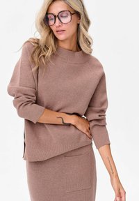 Jasnobrązowy sweter dzianinowy z wysokim kołnierzem i rękawami raglanowymi. Posiada luźny krój oraz lekko zaokrąglony dół po bokach.