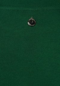 Grön ribbad tröja med en rund silverfärgad logotyp placerad nära halsringningen. Mjukt material med vertikala linjer. Strukturerad passform.