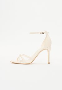 Beige high-heeled sandal with an ankle strap, featuring thin crisscrossing straps, a pointed toe, and a smooth, glossy texture.