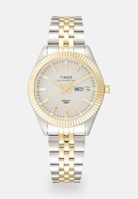 Timex ura s srebrno-zlatim nehrjajućim čeličnim remenom, okruglim srebrnim brojčanikom s oznakama sati, prikazom datuma i režnjem s rebrastim uzorkom.