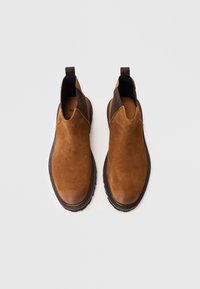 Botine din suede maron cu panouri elastice laterale și tab-uri pentru a fi trase, având o talpă texturată din cauciuc negru și un vârf neted.