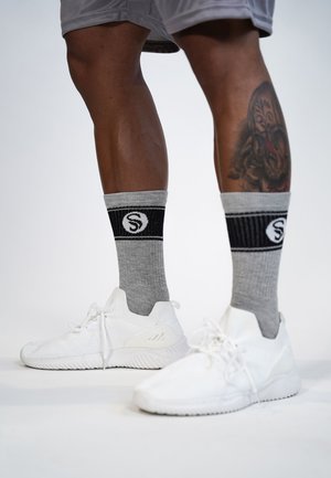 Weißer Sportschuhe mit strukturiertem Obermaterial und flexibler Sohle, kombiniert mit grauen und schwarzen, gerippten Socken, die ein kreisförmiges Logomuster aufweisen.