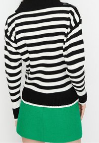 Sweter dziewiarski w czarno-białe paski z wysokim kołnierzem, o strukturze ribbed. W zestawieniu z kontrastującą zieloną spódnicą. Uzupełniony delikatnym białym wykończeniem.