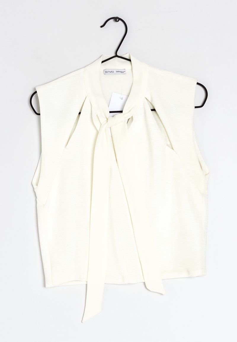 Mouwloze crèmekleurige top met een zachte, geribbelde textuur; heeft een centrale strik bij de kraag en openings aan de zijkanten. Hangt aan een zwarte haak.